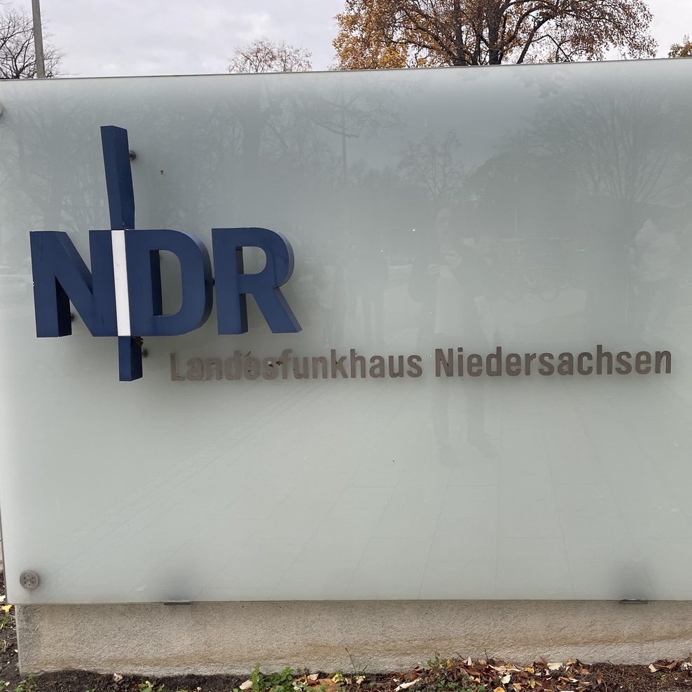 Auf einem großes weißes Hinweisschild vor dem Gebäude des NDRs steht in blauen Großbuchstaben "NDR", darunter in schwarzer, etwas kleinerer Schrift "Landesfunkhaus Niedersachsen"