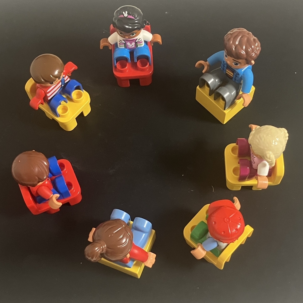 Auf dem Foto sind auf einer schwarzen Unterlage sieben Stühle aus Duplo-Steinen zu einem Sitzkreis angeordnet. Auf den Stühlen sitzen fünf kindliche Duplo-Männchen und zwei erwachsene Duplo-Männchen.