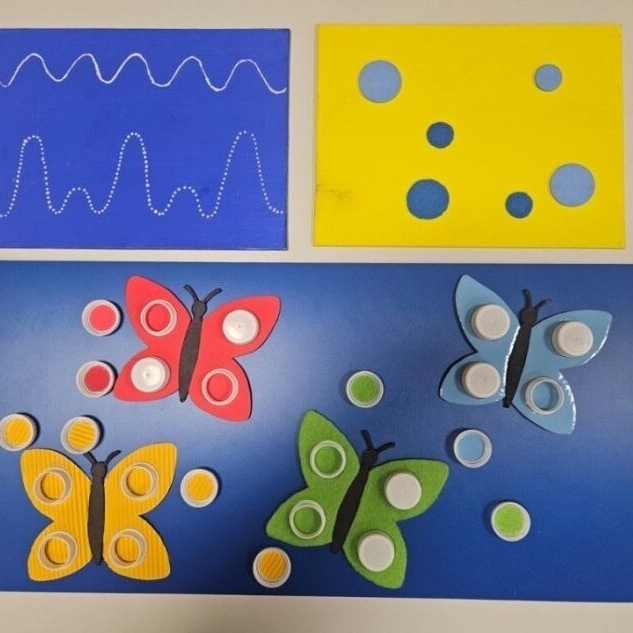 Auf dem Foto sind 3 Aufgabentafeln zu sehen. Auf einer blaue Aufgabentafel sind weiße tastbare Punkte zu sehen, die in Wellenform über die Tafel laufen. Auf einer gelben Tafel sind verschieden große und unterschiedlich tastbare Punkte in zwei Blautön