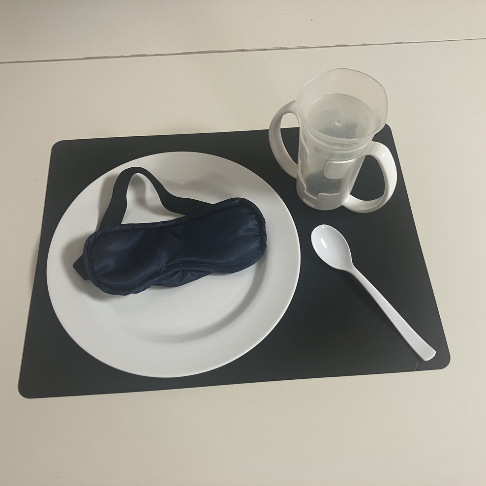 Auf einem schwarzen Set liegt links ein weißer Teller mit einer Schlafmaske darauf. Oben rechts steht ein Trinklernbecher mit Handgriffen. Unten rechts liegt ein weißer Löffel.