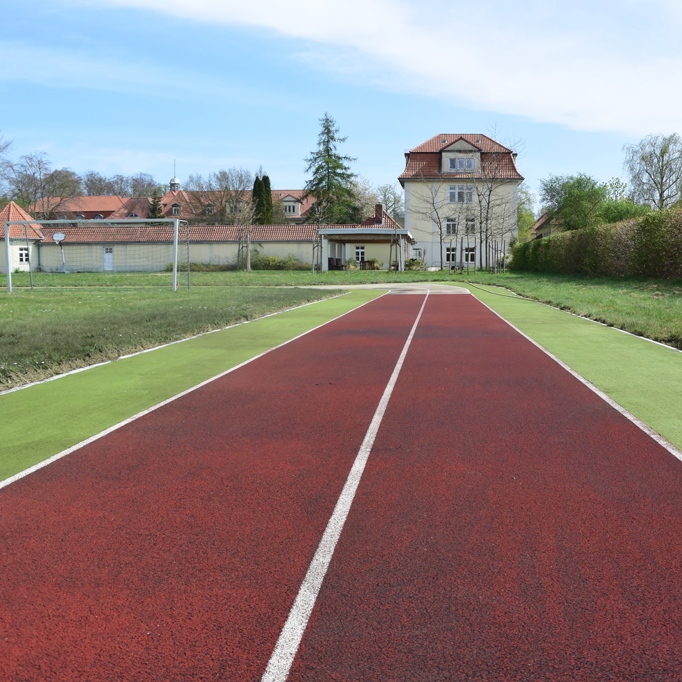 Blick auf eine Laufbahn/Tartanbahn mit einem Schulgebäude im Hintergrund.