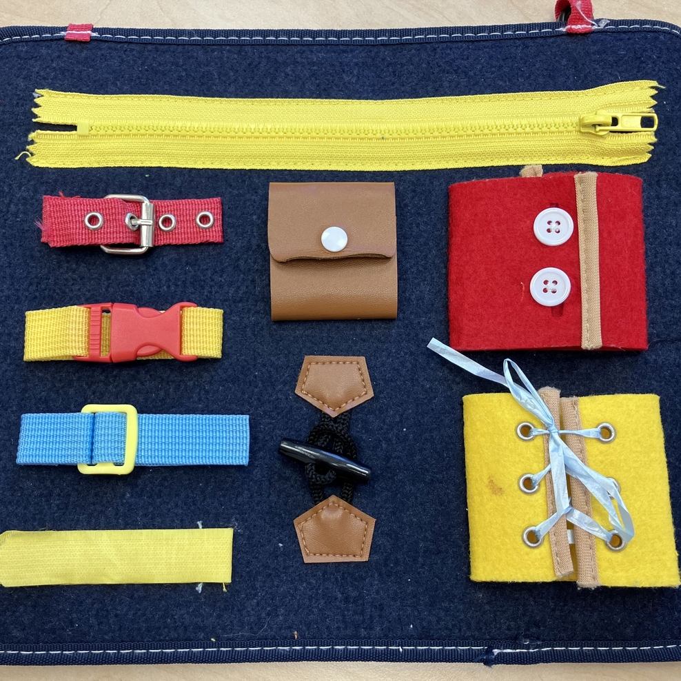 Das Bild zeigt ein Fördermaterial mit dem man lernen kann, Verschlüsse an Kleidung zu öff-nen und zu schließen. Auf einer Filzmatte sind Verschlüsse mit Knöpfen, Reißverschluss, Steckschnallen, Klett und zum Zubinden aufgenäht.