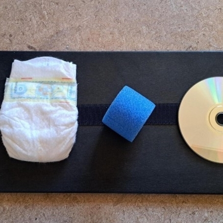 Auf einem schwarzen Holzbrett sind mit Klett drei Bezugsobjekte nebeneinander befestigt, links eine Windel, in der Mitte ein Stück Poolnudel und rechts eine CD.